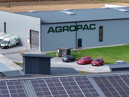 Halle mit AGROPAC- und ALC-Schriftzügen, geparkten Fahrzeugen und Solarmodulen im Vordergrund.