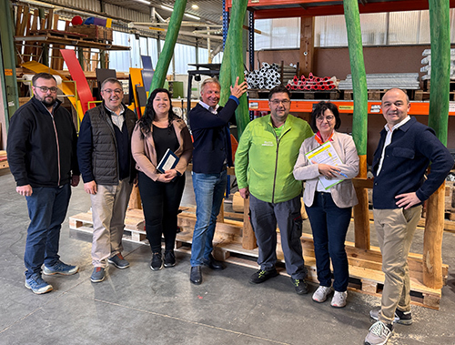 Sieben Personen vor aufgestellten grünen Holzstämmen in einer Lagerhalle.